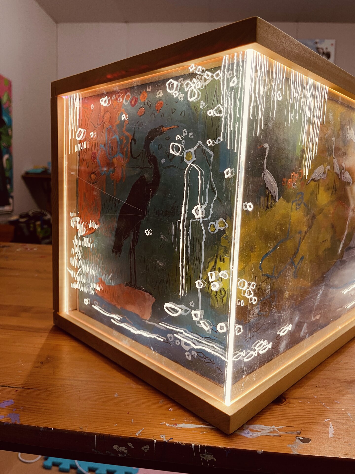 Angled view of a wooden-framed, box-like artwork with imagery across its front and left side. White long-legged birds stand among green and yellow forms above blue-grey water, while a dark bird silhouette appears on the left. Fine white lines and looping marks cross the scene.