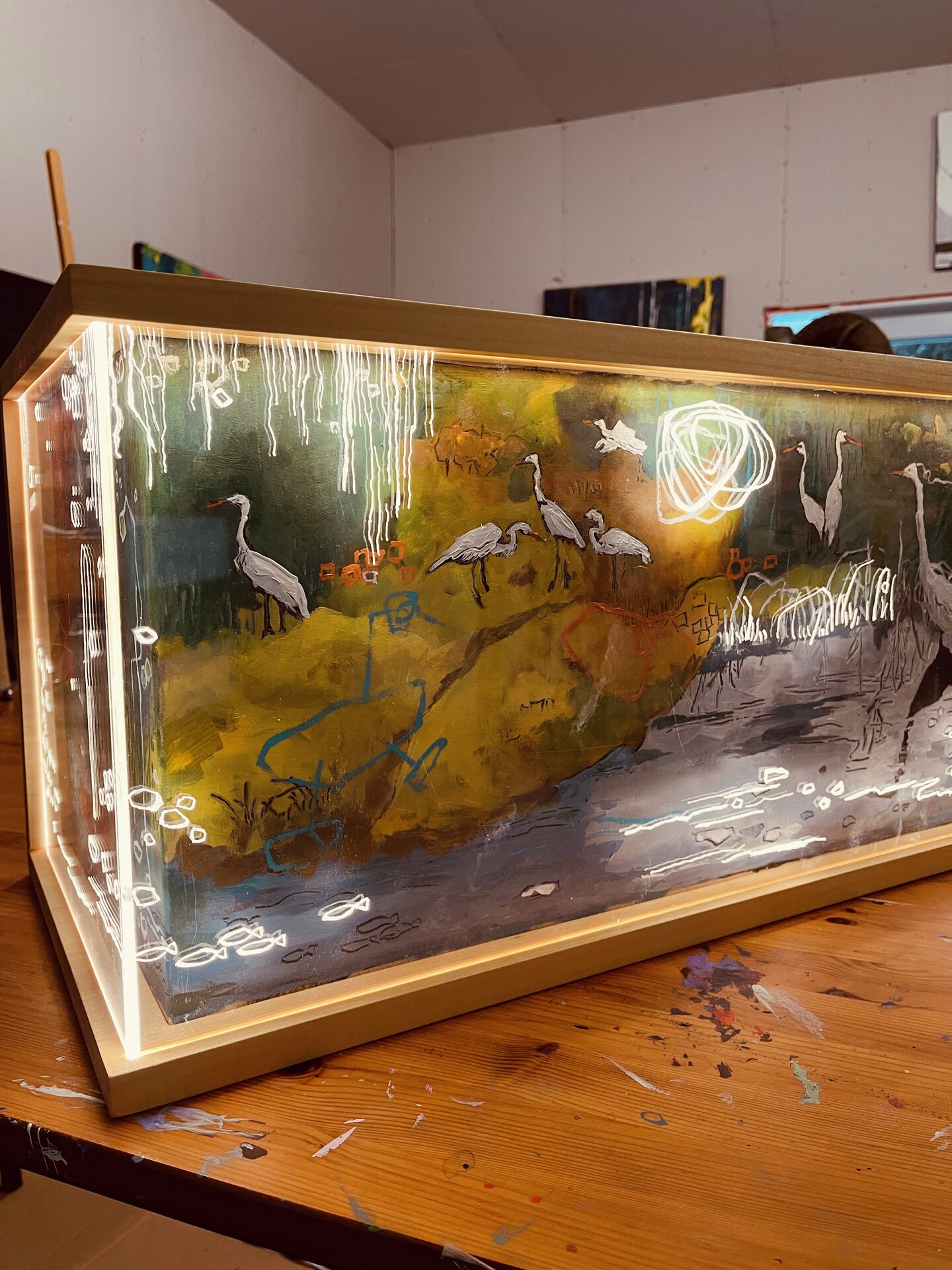 Angled view of a wooden-framed, box-like artwork with imagery across its front and left side. White long-legged birds stand among green and yellow forms above blue-grey water, while a dark bird silhouette appears on the left. Fine white lines and looping marks cross the scene.
