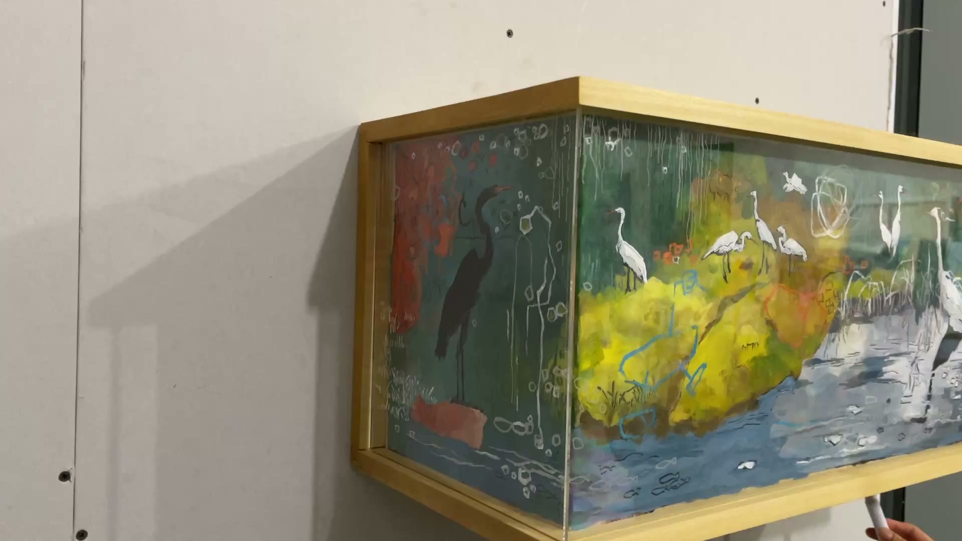 Angled view of a wooden-framed, box-like artwork with imagery across its front and left side. White long-legged birds stand among green and yellow forms above blue-grey water, while a dark bird silhouette appears on the left. Fine white lines and looping marks cross the scene.