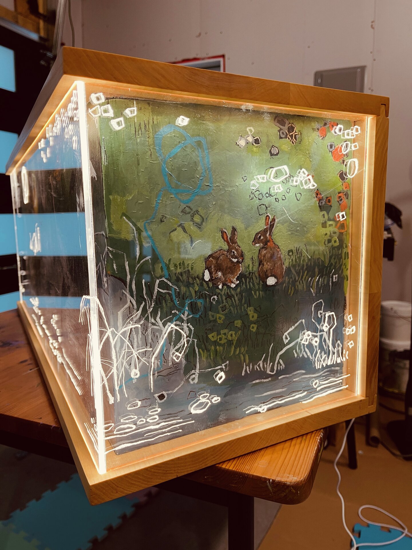 Angled view of a wooden-framed, box-like artwork with imagery across its front and left side. White long-legged birds stand among green and yellow forms above blue-grey water, while a dark bird silhouette appears on the left. Fine white lines and looping marks cross the scene.